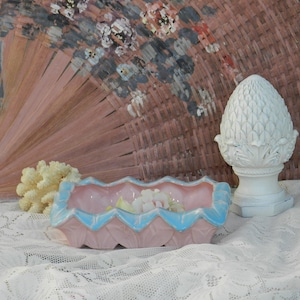 以下が含まれることがあります： ピンクとブルーのセラミック製の皿で、縁がギザギザになっています。皿は白いレースのテーブルクロスの上に置かれています。背景には白いセラミック製のパイナップルのフィニアルがあります。
