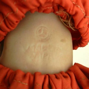 K&ouml;nnte beinhalten: Nahaufnahme eines Puppenkopfes mit hellem Teint, umrahmt von einem leuchtend orangefarbenen Stoff. Der Hals der Puppe tr&auml;gt eine gepr&auml;gte Markierung mit der Aufschrift "MACMA JAPAN". Der orangefarbene Stoff ist gerafft und gen&auml;ht.