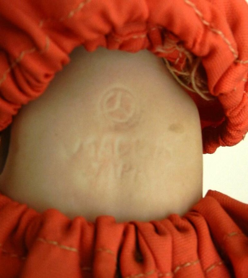 K&ouml;nnte beinhalten: Nahaufnahme eines Puppenkopfes mit hellem Teint, umrahmt von einem leuchtend orangefarbenen Stoff. Der Hals der Puppe tr&auml;gt eine gepr&auml;gte Markierung mit der Aufschrift "MACMA JAPAN". Der orangefarbene Stoff ist gerafft und gen&auml;ht.