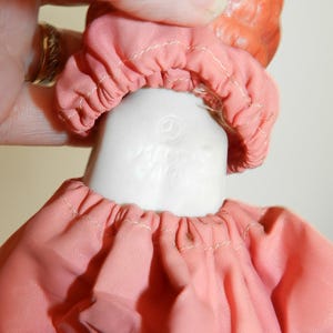 K&ouml;nnte beinhalten: Nahaufnahme von Kopf und Schultern einer Vintage-Puppe. Die Puppe hat einen pfirsichfarbenen Kopf und einen wei&szlig;en Porzellanhals. Sie tr&auml;gt ein rosa Stoffkleid mit gerafftem Ausschnitt und eine passende Haube. Die Worte "MADE IN JAPAN" sind sichtbar.