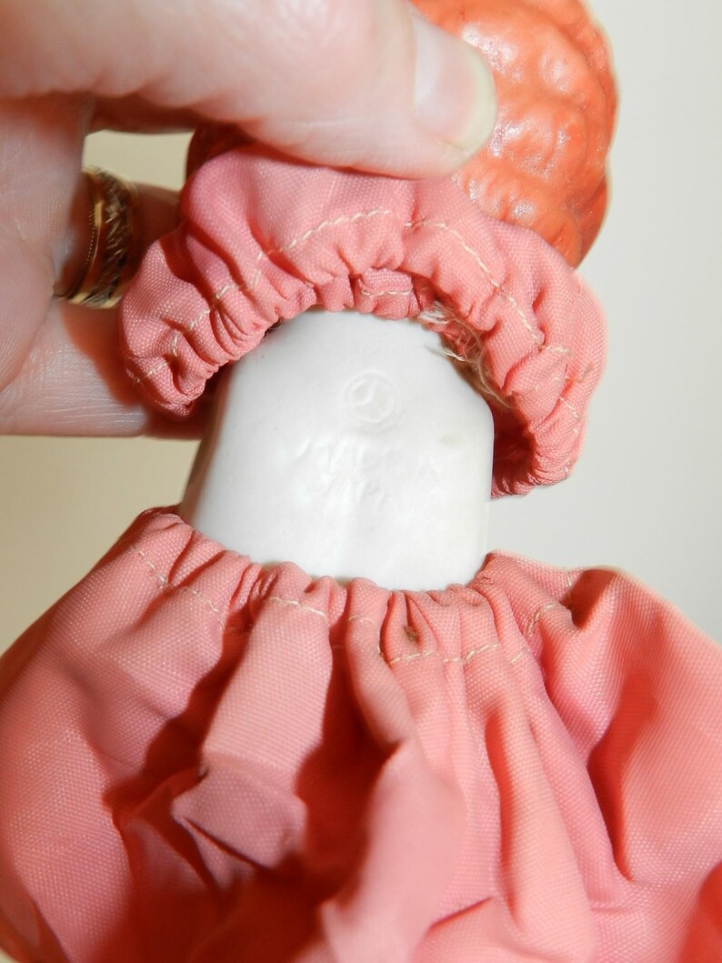 K&ouml;nnte beinhalten: Nahaufnahme von Kopf und Schultern einer Vintage-Puppe. Die Puppe hat einen pfirsichfarbenen Kopf und einen wei&szlig;en Porzellanhals. Sie tr&auml;gt ein rosa Stoffkleid mit gerafftem Ausschnitt und eine passende Haube. Die Worte "MADE IN JAPAN" sind sichtbar.