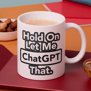 May include: A white ceramic mug featuring the black text "Hold On Let Me ChatGPT That." The mug has a standard handle and is filled with a light brown beverage. The design includes a rectangular border around the word ChatGPT.
