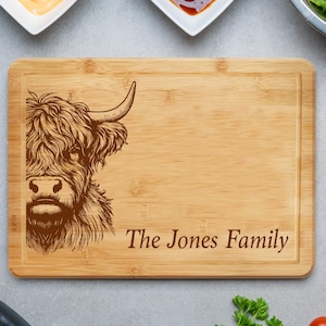 May include: A rectangular bamboo cutting board with a carved image of a Highland cow and the text "The Jones Family". The board is surrounded by small bowls of sauces, a salad, a hamburger bun, tomatoes, and parsley.
