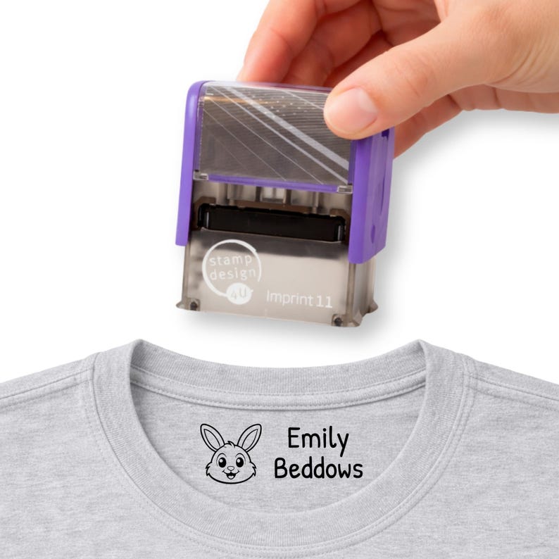 May include: A purple and silver self-inking stamp is held above a gray t-shirt. The stamp is labeled "stamp design" and "Imprint 11". The t-shirt has a bunny graphic and the name "Emily Beddows" printed on it.