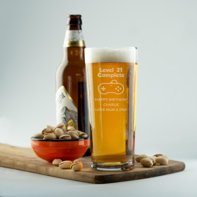 Op de afbeelding: Een pintglas gevuld met bier, met de tekst "Level 21 Complete" en een game controller-ontwerp. Het glas bevat ook de tekst "Happy Birthday Charlie Love Mum & Dad". Een bierfles en een kom pistachenoten zijn ook zichtbaar.