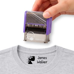 May include: A purple and silver stamp with the text "stamp design" and "Imprint 11" is held above a gray t-shirt. The t-shirt has a black dinosaur graphic and the name "James Walker" printed on it.
