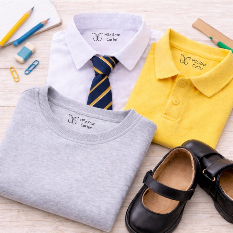 May include: School uniform items including a white collared shirt with a blue and gold striped tie, a gray sweatshirt, and a yellow polo shirt. Each item has a label with a butterfly graphic and the text "Mila Rose Carter". Black Mary Jane shoes are also present.