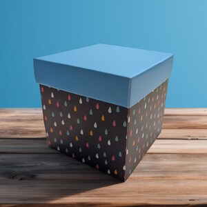 Puede incluir: Una caja de regalo cuadrada con tapa azul claro y base negra. La base est&aacute; decorada con un patr&oacute;n de gotas de colores. La caja est&aacute; sobre una superficie de madera.