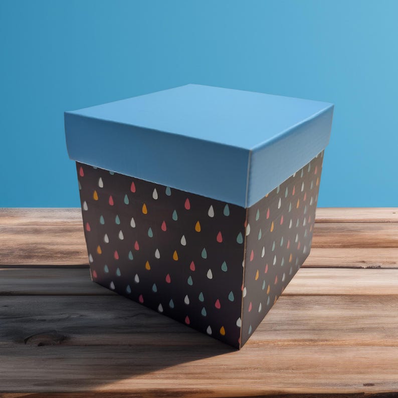 Puede incluir: Una caja de regalo cuadrada con tapa azul claro y base negra. La base est&aacute; decorada con un patr&oacute;n de gotas de colores. La caja est&aacute; sobre una superficie de madera.