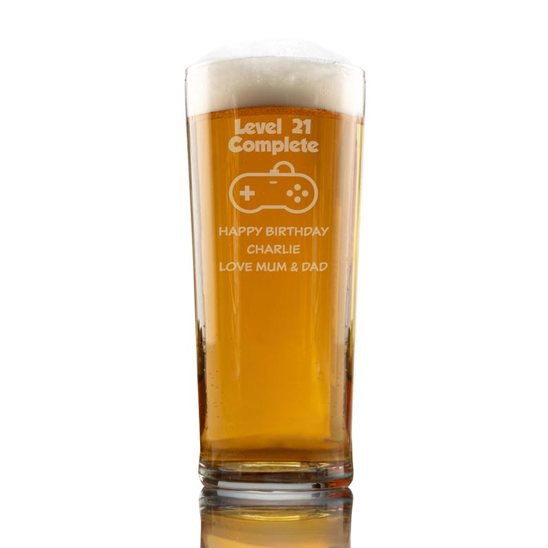 Op de afbeelding: Een pintglas gevuld met bier en schuim. Het glas heeft de tekst "Level 21 Complete" met een gamecontroller-afbeelding en "HAPPY BIRTHDAY CHARLIE LOVE MUM & DAD" gegraveerd. Het glas is helder en staat op een witte ondergrond.