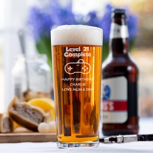 Op de afbeelding: Een hoog, helder glas gevuld met bier en schuim, met de tekst "Level 21 Complete" en een gamecontroller-afbeelding. Daaronder staat "Happy Birthday Charlie, Love Mum & Dad". Op de achtergrond een bruine bierfles.