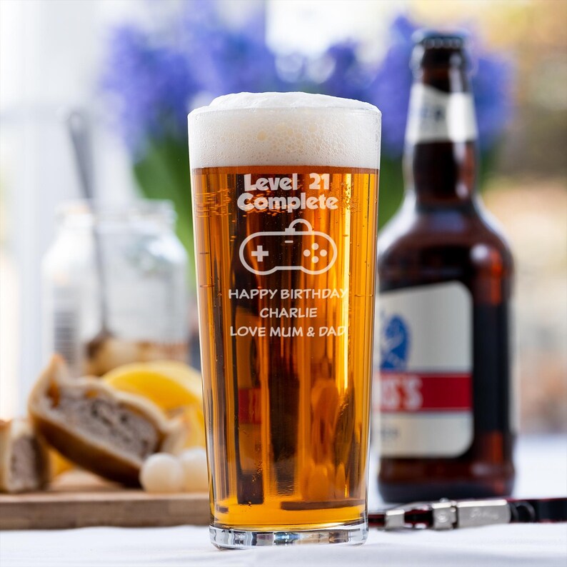 Op de afbeelding: Een hoog, helder glas gevuld met bier en schuim, met de tekst "Level 21 Complete" en een gamecontroller-afbeelding. Daaronder staat "Happy Birthday Charlie, Love Mum & Dad". Op de achtergrond een bruine bierfles.