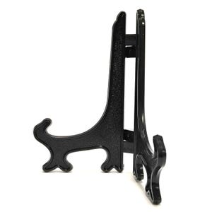 May include: A black plastic display stand featuring a hinged design with two connected arms. The stand has a textured surface and curved, decorative feet at the base of each arm, designed to hold items upright in a stable, open position.