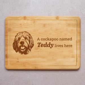 May include: A rectangular bamboo chopping board with a carved border. It features a detailed illustration of a Cockapoo dog and the text "A cockapoo named Teddy lives here" in brown.