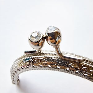 Puede incluir: Cierre de bolso plateado con detalles ornamentados y dos acentos de cristal transparente.