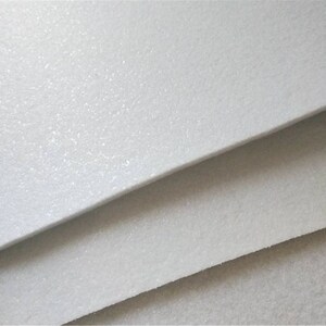 May include: Close-up of a white, textured, felt-like material. The material is layered in a stack, showing the texture and thickness.
