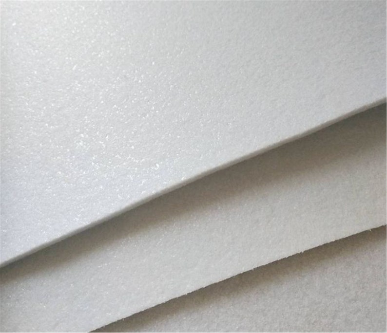 May include: Close-up of a white, textured, felt-like material. The material is layered in a stack, showing the texture and thickness.