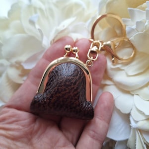 May include: A small, brown leather coin purse with a gold-colored frame and clasp, attached to a gold keychain. The purse has a textured surface and is set against a backdrop of white flowers.
