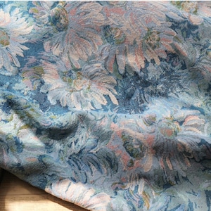 May include: A woven fabric featuring a dense floral pattern in shades of blue, white, and soft pink. The textile displays a textured, tapestry-like appearance with overlapping daisy-like flower motifs across the entire surface of the material.