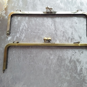May include: Two metal purse frames in silver and brass tones. The frames are rectangular with rounded corners and a clasp closure. The silver frame has a shiny finish, while the brass frame has a matte finish. Both have small loops for attaching to a purse.