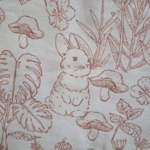 May include: Off-white fabric with a repeating pattern of brown line drawings. The design includes a rabbit, mushrooms, flowers, and various plants. The overall theme is a whimsical woodland scene.