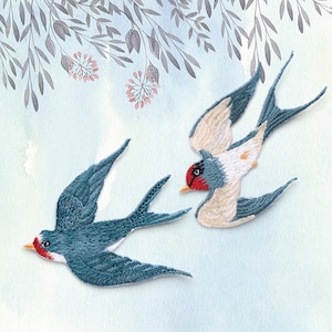 Peut inclure: Deux hirondelles brodées avec des accents bleus, blancs et rouges. Les oiseaux volent dans des directions opposées sur un fond bleu clair avec des branches grises de style aquarelle et des fleurs roses.