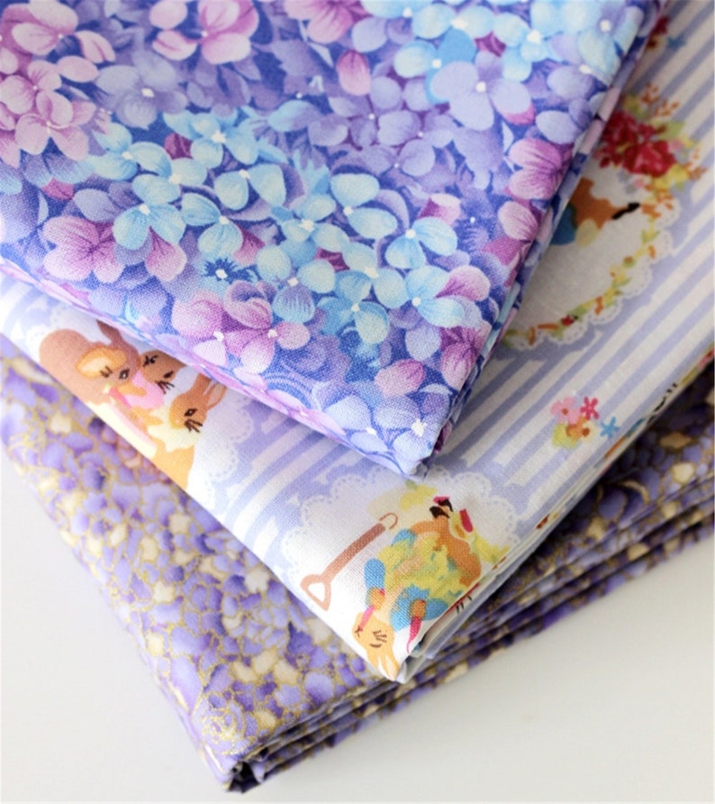 May include: A stack of three fabric squares with different floral and patterned designs. The top square features a purple and blue floral pattern, the middle square has a white and blue striped pattern with small floral accents, and the bottom square has a purple and gold speckled pattern.