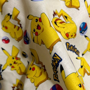 May include: Cream-coloured fabric with a repeating pattern of yellow Pikachu characters, red and blue Pokeball designs, and the word "Pokemon" in blue. The fabric has a soft, slightly wrinkled texture.