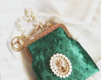 May include: A dark green velvet pouch with a gold-toned metal kiss-lock frame. The front features an oval cameo brooch surrounded by white faux pearls. A short handle made of white faux pearls and a gold-toned metal lobster clasp are attached.