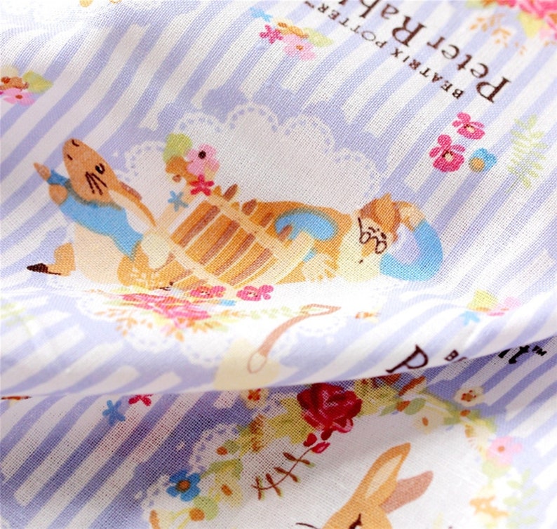 May include: A white fabric with a blue and white striped pattern. The fabric features a colorful illustration of Peter Rabbit and a teapot with flowers and text that reads "Beatrix Potter Peter Rabbit".