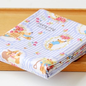 May include: A blue and white striped fabric with a repeating pattern of Peter Rabbit characters and floral designs. The fabric features the text "Peter Rabbit" and "Beatrix Potter".