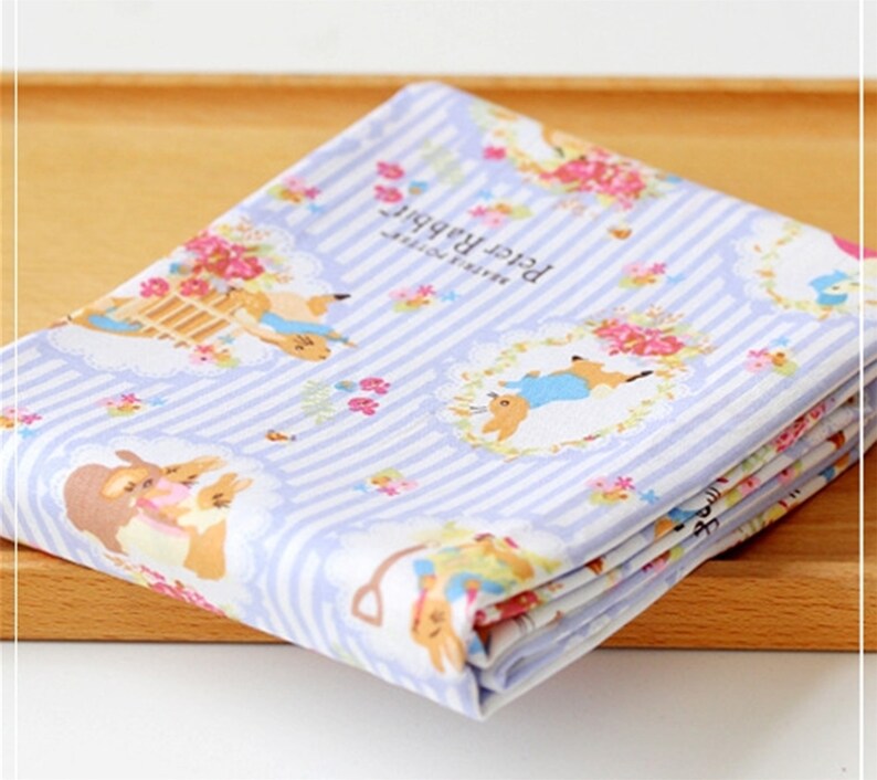 May include: A blue and white striped fabric with a repeating pattern of Peter Rabbit characters and floral designs. The fabric features the text "Peter Rabbit" and "Beatrix Potter".