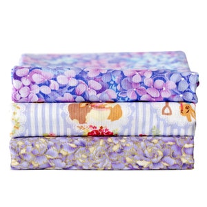 May include: A stack of three fabric squares with different floral patterns. The top square is purple with a floral pattern, the middle square is light blue with a striped pattern, and the bottom square is a light purple with a floral pattern.