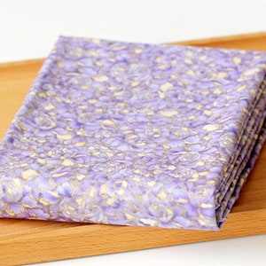 May include: Purple fabric with a floral pattern featuring gold accents. The fabric is folded and displayed on a wooden surface.