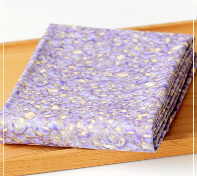 May include: Purple fabric with a floral pattern featuring gold accents. The fabric is folded and displayed on a wooden surface.