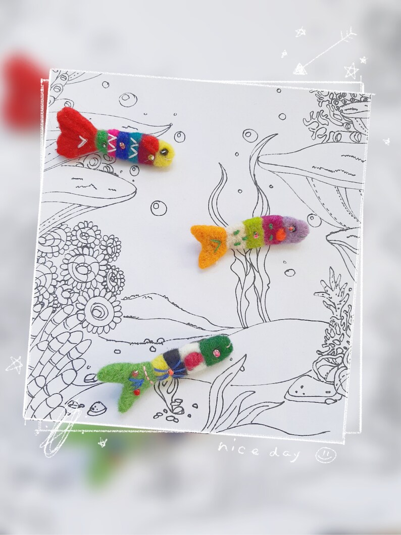 May include: Three small, handcrafted felt fish ornaments in vibrant, multicolored patterns with embroidered details and bead accents, displayed on a white card featuring a black and white line drawing of an underwater coral reef scene.