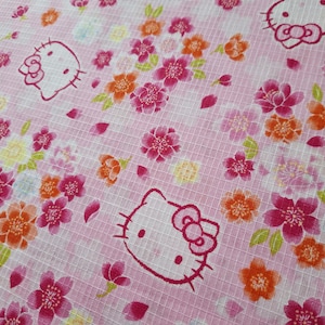 Puede incluir: Tela rosa con un patrón repetido de caras de Hello Kitty y flores coloridas. Las flores son en tonos de rosa, naranja y amarillo, con hojas verdes. La tela tiene una textura de rejilla.