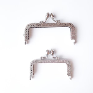 Puede incluir: Dos marcos rectangulares para bolso de metal plateado con mecanismo de cierre de boquilla. Cada marco presenta un intrincado patrón floral en relieve en los bordes exteriores y dos perillas redondeadas en la parte superior para abrir.