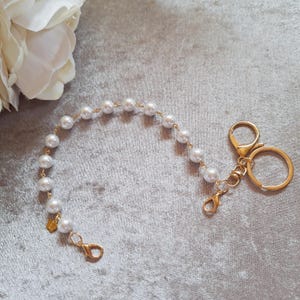May include: A gold-coloured keychain featuring a pearl beaded chain. The chain is made of small, round, white pearls strung together with gold wire. The keychain has a lobster clasp and a key ring.