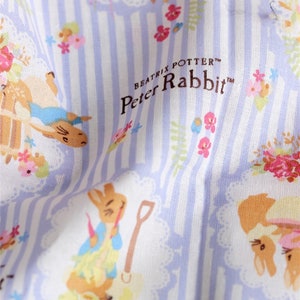 May include: White fabric with a blue and white striped pattern. The fabric features a repeating pattern of Peter Rabbit characters and floral designs. The text "Beatrix Potter&trade; Peter Rabbit&trade;" is printed in brown on the fabric.