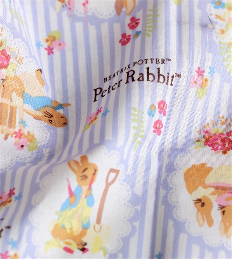 May include: White fabric with a blue and white striped pattern. The fabric features a repeating pattern of Peter Rabbit characters and floral designs. The text "Beatrix Potter&trade; Peter Rabbit&trade;" is printed in brown on the fabric.