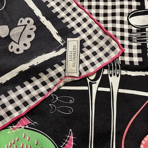 May include: A black and white silk scarf with a pink trim. The scarf displays a gingham pattern, fish, and cutlery illustrations. The label reads "Lulu Guinness".