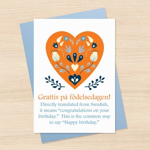 May include: A greeting card with a blue envelope. The card features a large orange heart with floral designs in white, blue, and navy. The text reads "Grattis på födelsedagen!" which means "Happy birthday."