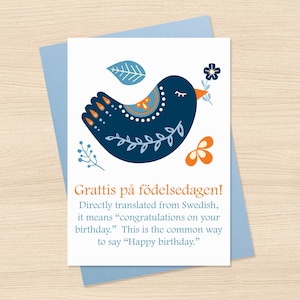 May include: A greeting card with a blue envelope. The card features a stylized blue bird with orange and white accents, holding a flower. The text "Grattis på födelsedagen!" is displayed, meaning "Happy birthday."