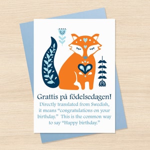 May include: A greeting card with a stylized orange fox illustration, blue accents, and the text "Grattis på födelsedagen!" which translates to "Happy birthday." The card is on a light blue envelope.