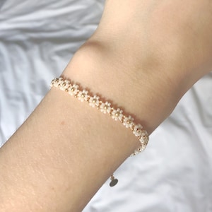 Könnte beinhalten: Ein zartes Armband mit einem Gänseblümchen-Ketten-Design. Das Armband besteht aus kleinen weißen Perlen und goldfarbenen Perlen. Das Armband wird am Handgelenk getragen und hat einen kleinen herzförmigen Anhänger.