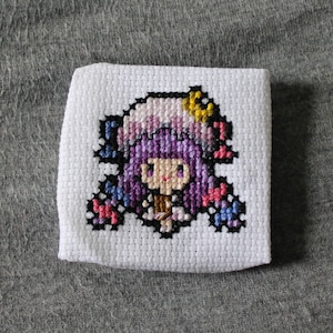 May include: A small, square cross-stitch embroidery on white fabric. The design features a cartoon character with purple hair, a yellow hair accessory, and a pink and blue outfit. The embroidery is detailed with black outlines and various colors.