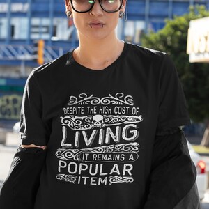 May include: Black t-shirt with a white graphic that says "Despite the high cost of living it remains a popular item" with a skull graphic above the word "living".
