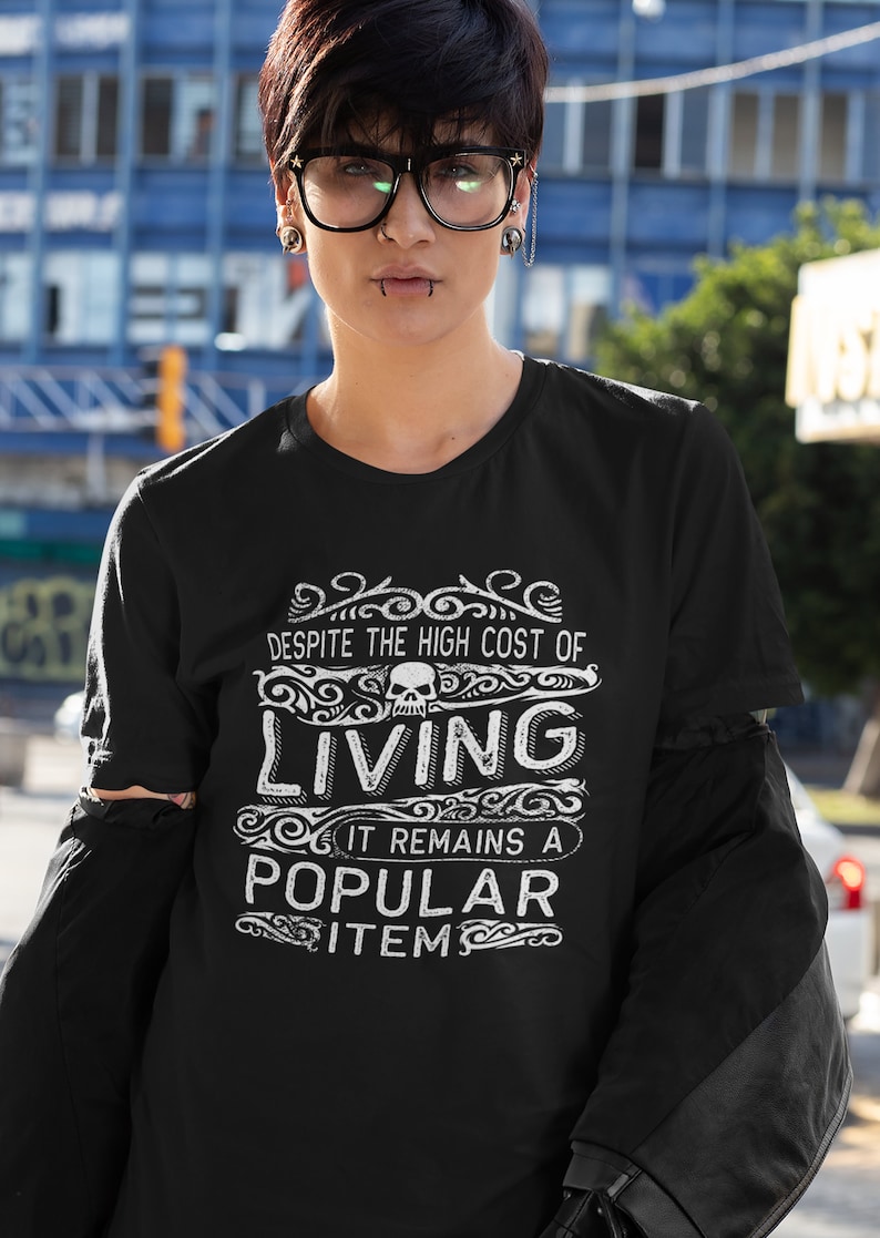 May include: Black t-shirt with a white graphic that says "Despite the high cost of living it remains a popular item" with a skull graphic above the word "living".