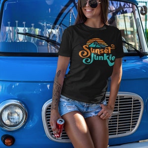 May include: A woman wearing a black t-shirt with a sunset graphic and the text "Sunset Junkie" in a retro font. She is standing in front of a blue van and holding a can of soda.
